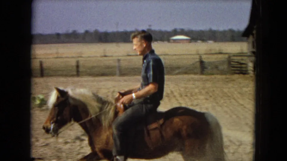The rider continues across the farmyard on the small pony, with open fields, fence posts, and a low distant building visible in the background.