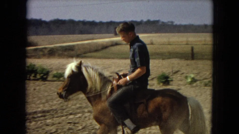 A closer home-movie frame shows the man seated on the small brown pony, holding the reins as the farm field and distant tree line stretch behind him.