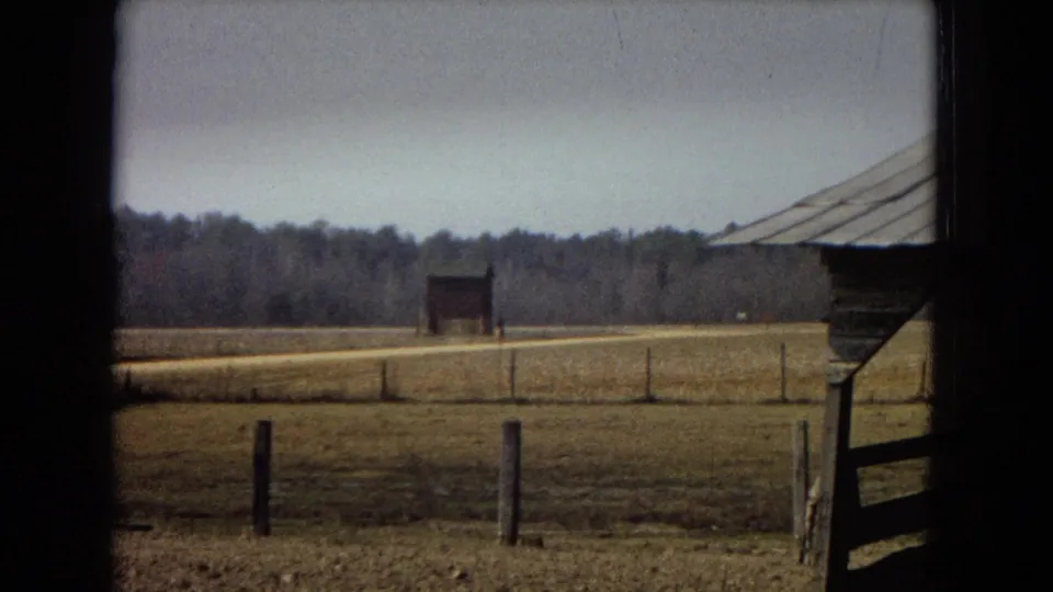 1967 archival 8mm home-movie frame looking from shade toward an open rural field, fence posts, a pale road, treeline, and a small distant barn.