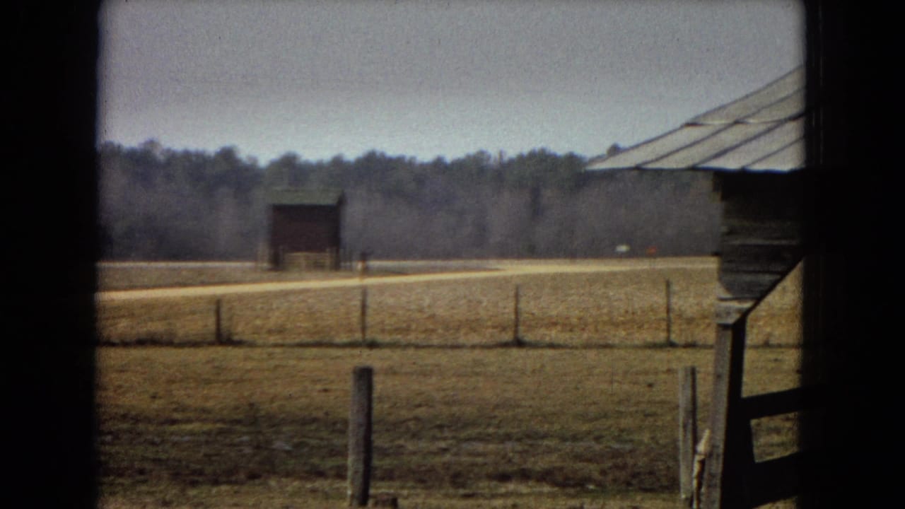 Open field with fence posts, a distant barn, treeline, and the edge of a wooden structure at right.