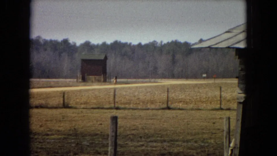 1967 archival 8mm frame showing an open rural field, a simple fence line, a small red barn near a dirt road, and a treeline under a pale sky.