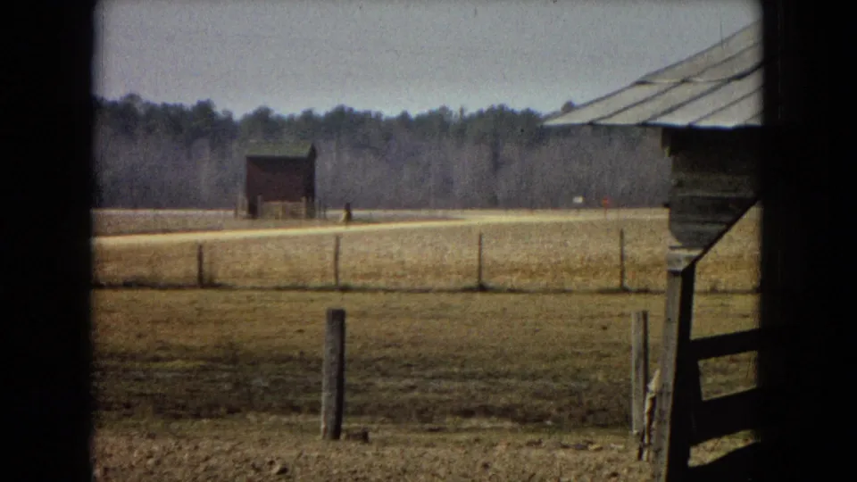 1967 archival 8mm home-movie frame of a rural field with foreground fence posts, a small barn, distant tiny figures near the road, and a wooded horizon.