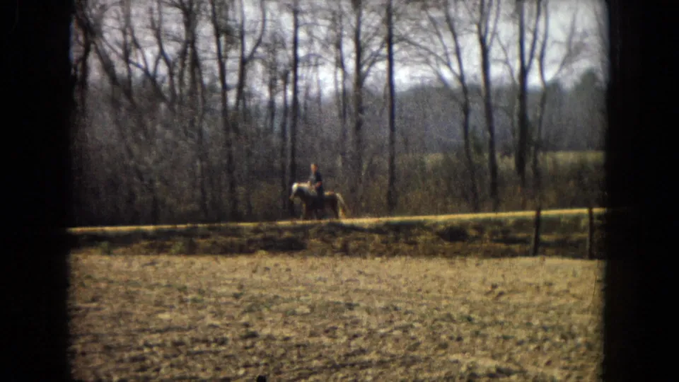 1967 U.S. 8mm home-movie frame with a man riding a light-colored horse along a sunlit rural path, seen through a dark film border.