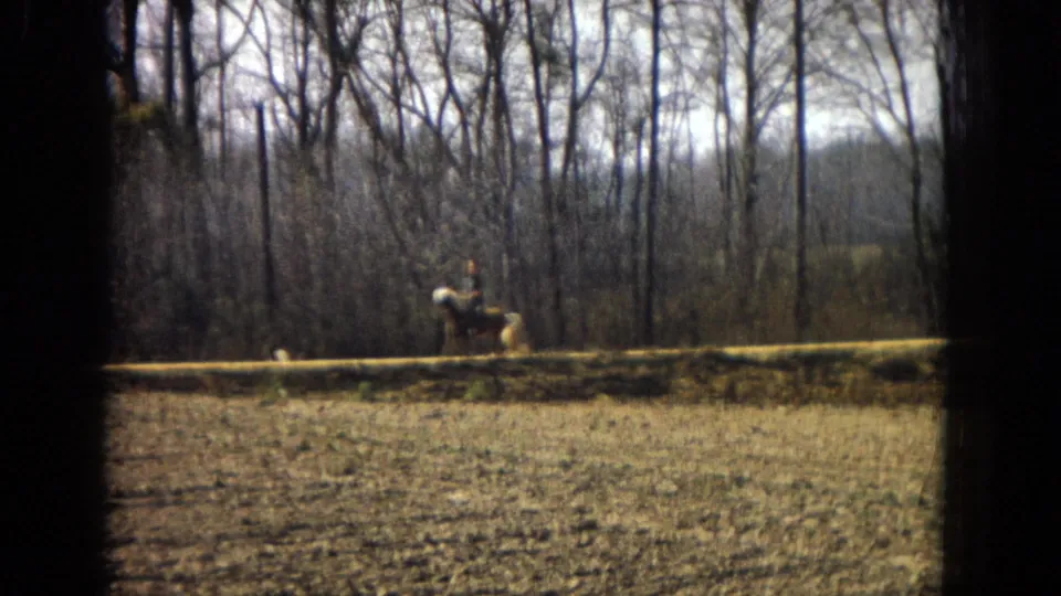 1967 U.S. 8mm home-movie frame showing the rider and light-colored horse moving past small pine saplings, bare trees, dry grass, and a low fence.
