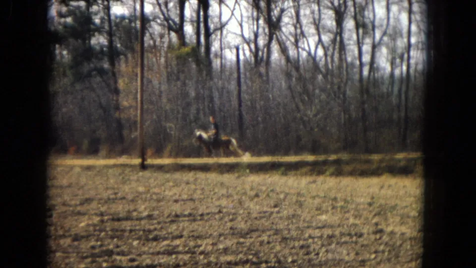 1967 U.S. 8mm home-movie frame of a distant man on a light-colored horse traveling along a rural path with dry grass, fencing, and leafless trees behind him.