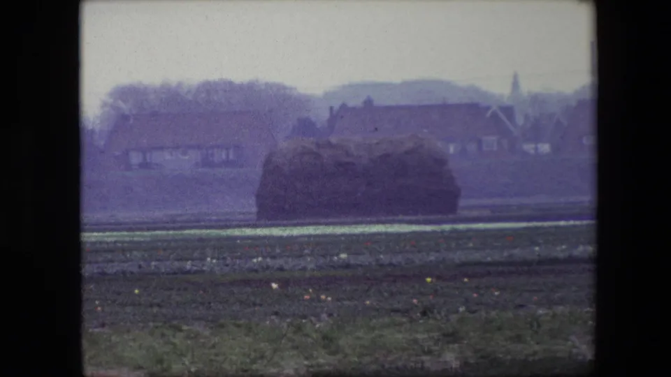1967 Brussels-area 8mm frame showing clustered hay bales in a field with distant houses, bare trees, low hills, and a small spire visible on the horizon.