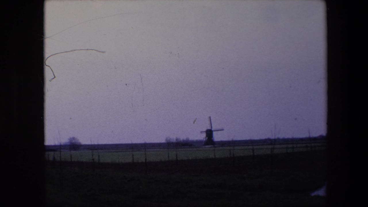 1967 Brussels-area 8mm frame with a distant traditional windmill silhouetted on the horizon beyond a flat field and fence line.
