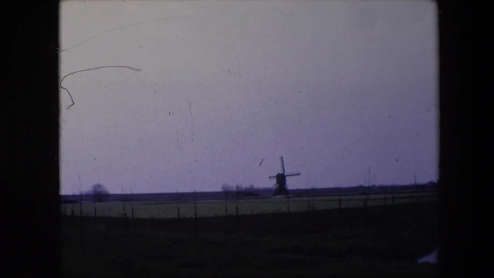 1967 Brussels-area 8mm frame with a distant traditional windmill silhouetted on the horizon beyond a flat field and fence line.