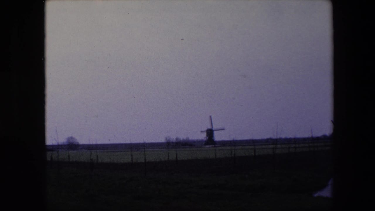 1967 Brussels-area home-movie view of a small windmill across open farmland, with fence posts and a muted cloudy sky.