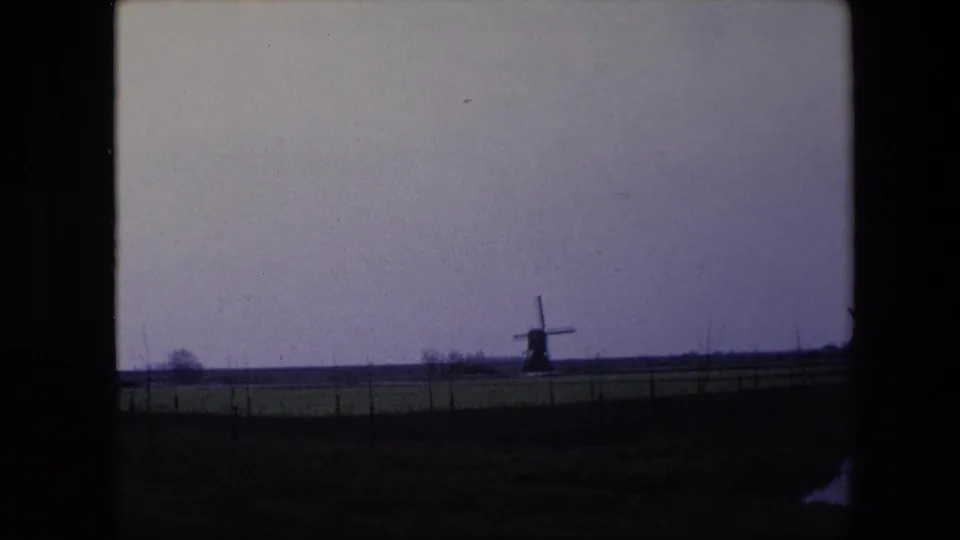 1967 Brussels-area home-movie view of a small windmill across open farmland, with fence posts and a muted cloudy sky.