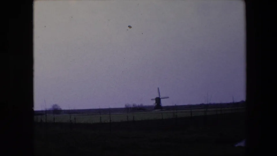 1967 Brussels-area 8mm frame showing a distant windmill in a flat rural landscape, framed by fence lines, cloudy sky, and dark film edges.