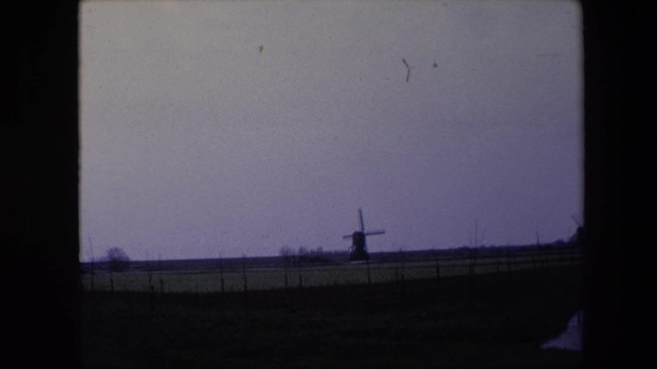 1967 8mm home-movie frame from the Brussels, Belgium metadata showing a distant windmill across a flat fenced pasture beneath a muted purple-gray sky.