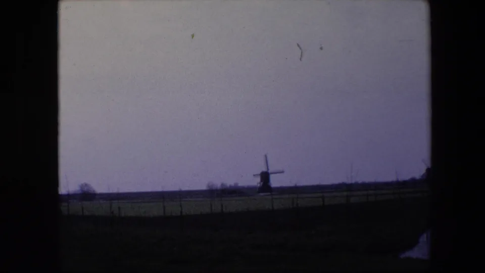 1967 8mm home-movie frame from the Brussels, Belgium metadata showing a distant windmill across a flat fenced pasture beneath a muted purple-gray sky.