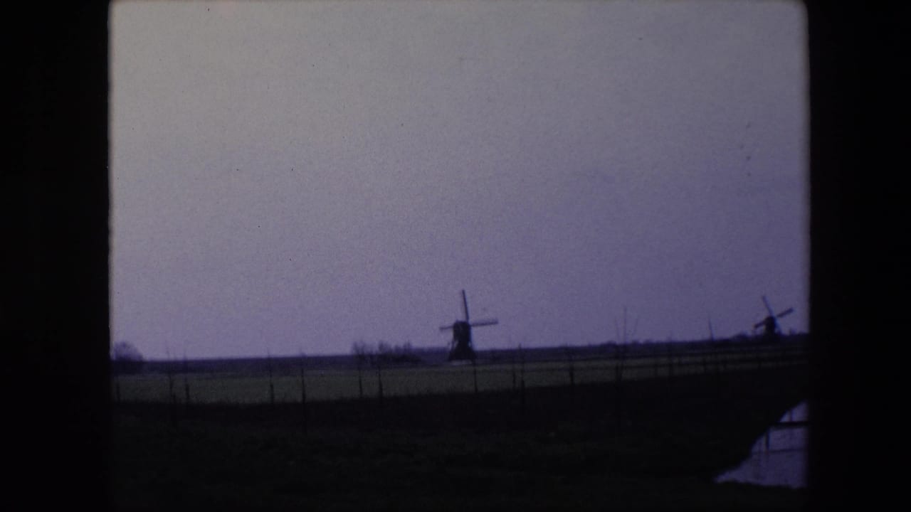 1967 8mm home-movie frame from the Brussels, Belgium metadata showing two windmills on the horizon beyond fenced fields and dark frame edges.
