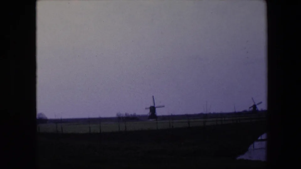 1967 8mm home-movie frame from the Brussels, Belgium metadata showing two windmills on the horizon beyond fenced fields and dark frame edges.