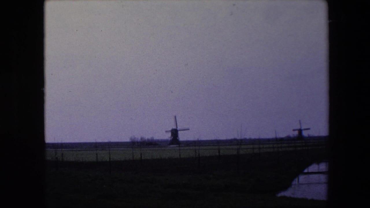 1967 8mm home-movie frame from the Brussels, Belgium metadata showing distant windmills across a fenced pasture, with a small water edge visible in the lower right.