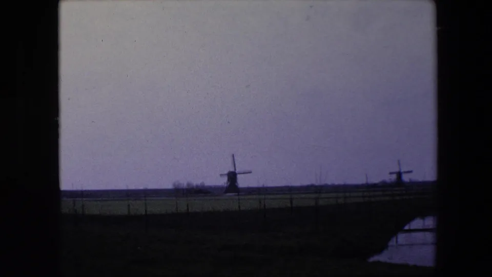 1967 8mm home-movie frame from the Brussels, Belgium metadata showing distant windmills across a fenced pasture, with a small water edge visible in the lower right.