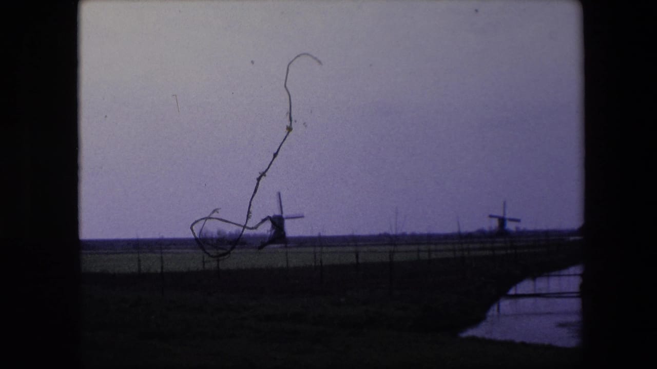 1967 8mm home-movie frame from the Brussels, Belgium metadata showing windmills across a flat fenced field, with a prominent film scratch or debris mark over the sky.