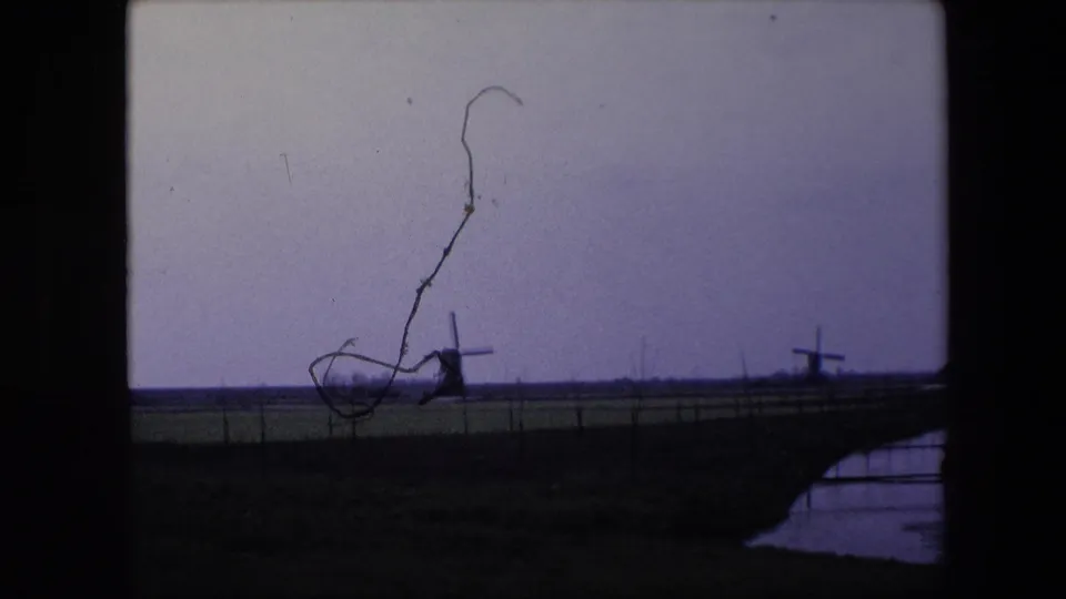 1967 8mm home-movie frame from the Brussels, Belgium metadata showing windmills across a flat fenced field, with a prominent film scratch or debris mark over the sky.