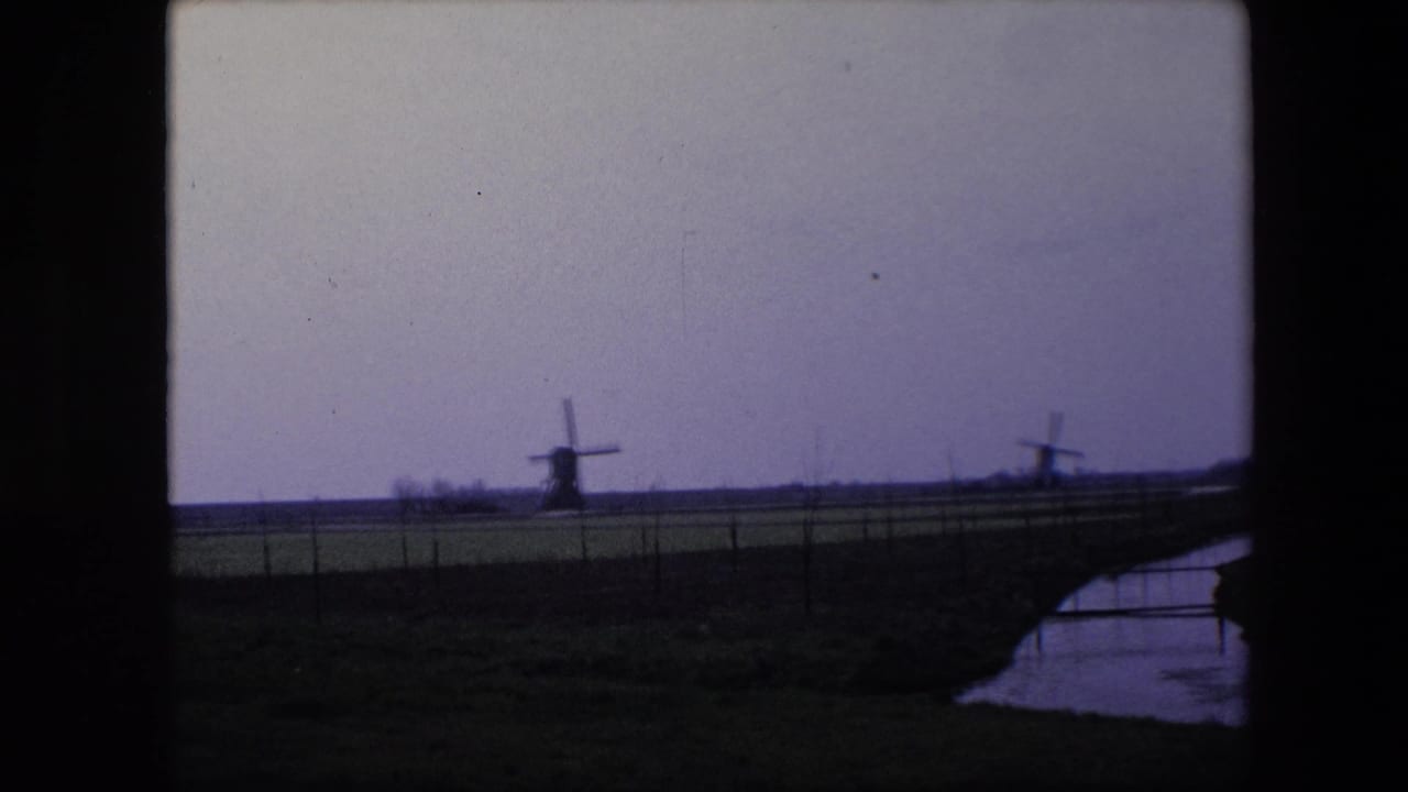 1967 8mm home-movie frame from the Brussels, Belgium metadata showing two distant traditional windmills, flat fields, fencing, and a narrow creek or canal at right.