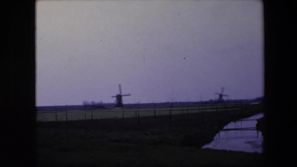 1967 8mm home-movie frame from the Brussels, Belgium metadata showing two distant traditional windmills, flat fields, fencing, and a narrow creek or canal at right.