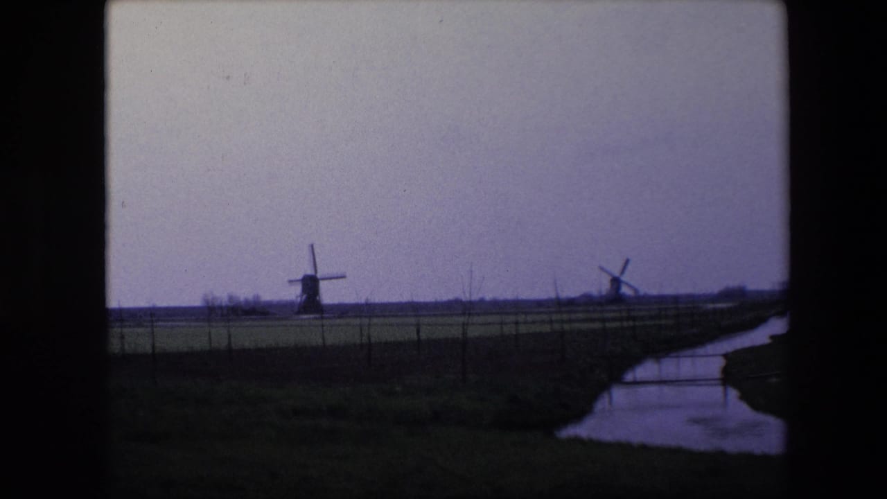 1967 Brussels-area home-movie frame showing two windmills on the horizon, a low fenced field, and a reflective creek or canal running along the right edge.