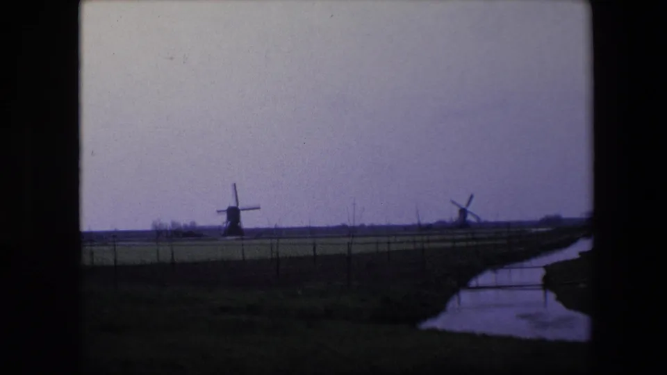 1967 Brussels-area home-movie frame showing two windmills on the horizon, a low fenced field, and a reflective creek or canal running along the right edge.