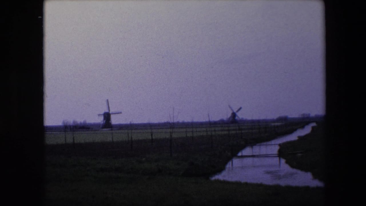 1967 8mm frame from the Brussels, Belgium clip showing distant windmills beyond fenced open land, with a narrow water channel and dark film-frame edges.