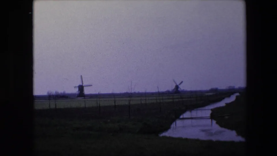 1967 8mm frame from the Brussels, Belgium clip showing distant windmills beyond fenced open land, with a narrow water channel and dark film-frame edges.