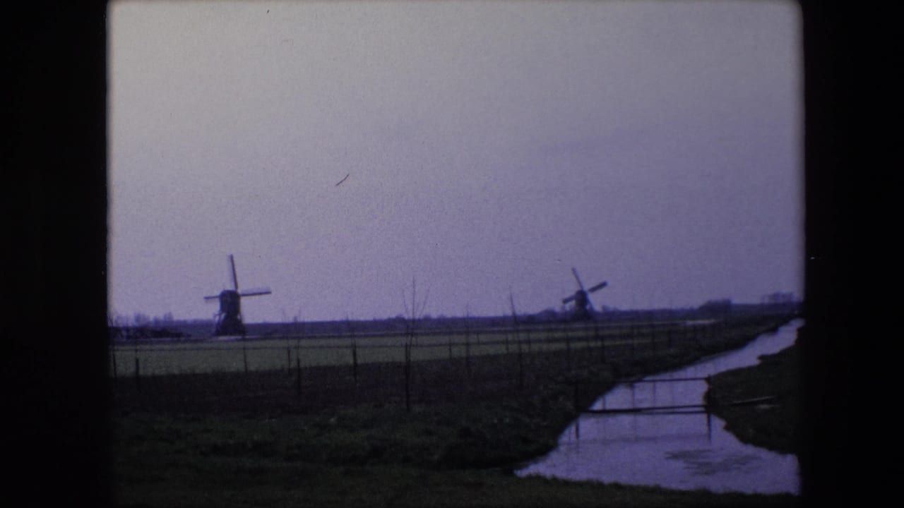 1967 Brussels, Belgium home-movie frame with two traditional windmills in a flat field landscape, a curving canal or creek, fence posts, and muted blue-purple film color.