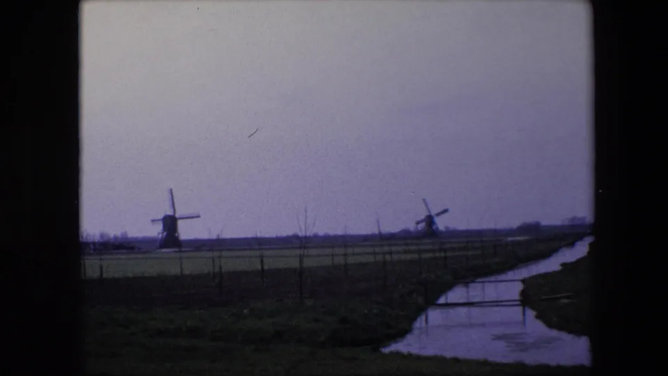 1967 Brussels, Belgium home-movie frame with two traditional windmills in a flat field landscape, a curving canal or creek, fence posts, and muted blue-purple film color.