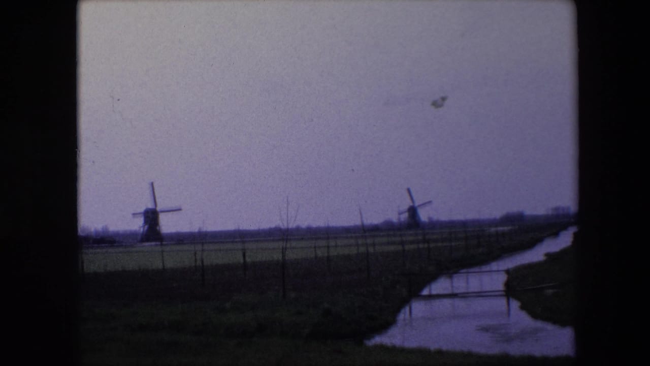 1967 Brussels-area home-movie frame showing two distant windmills across flat farmland, with a narrow canal or ditch and fence line in the foreground.