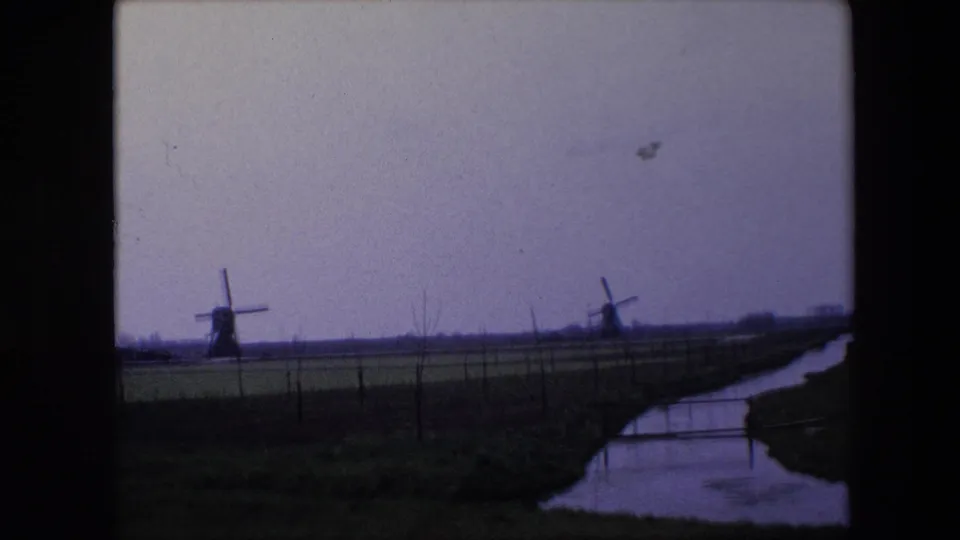 1967 Brussels-area home-movie frame showing two distant windmills across flat farmland, with a narrow canal or ditch and fence line in the foreground.