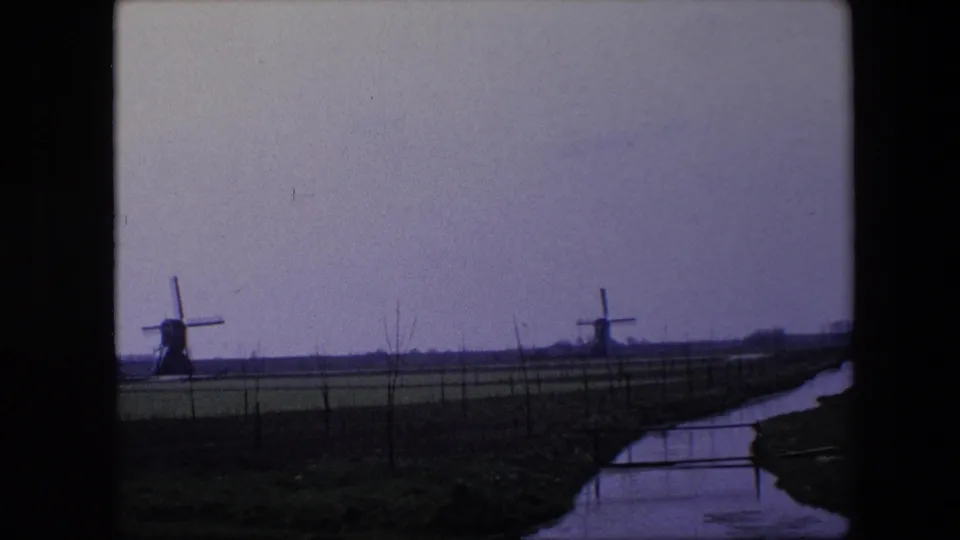 1967 Belgium countryside frame with two traditional windmills in the distance, a canal or drainage ditch at right, and sparse fence lines across the field.