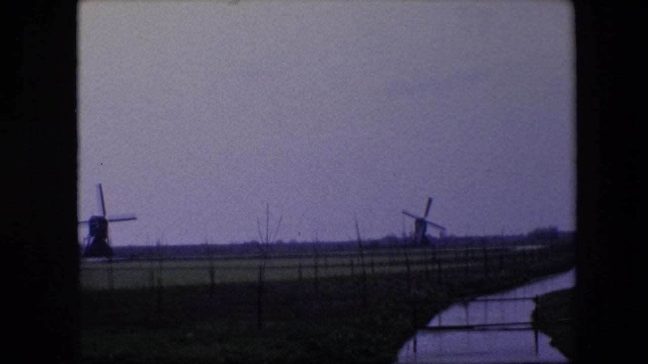 Soft 8mm frame from the 1967 Brussels-area clip showing distant windmills over open farmland, with a canal or ditch reflecting the gray sky.