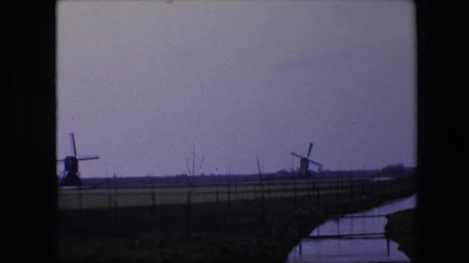 Soft 8mm frame from the 1967 Brussels-area clip showing distant windmills over open farmland, with a canal or ditch reflecting the gray sky.