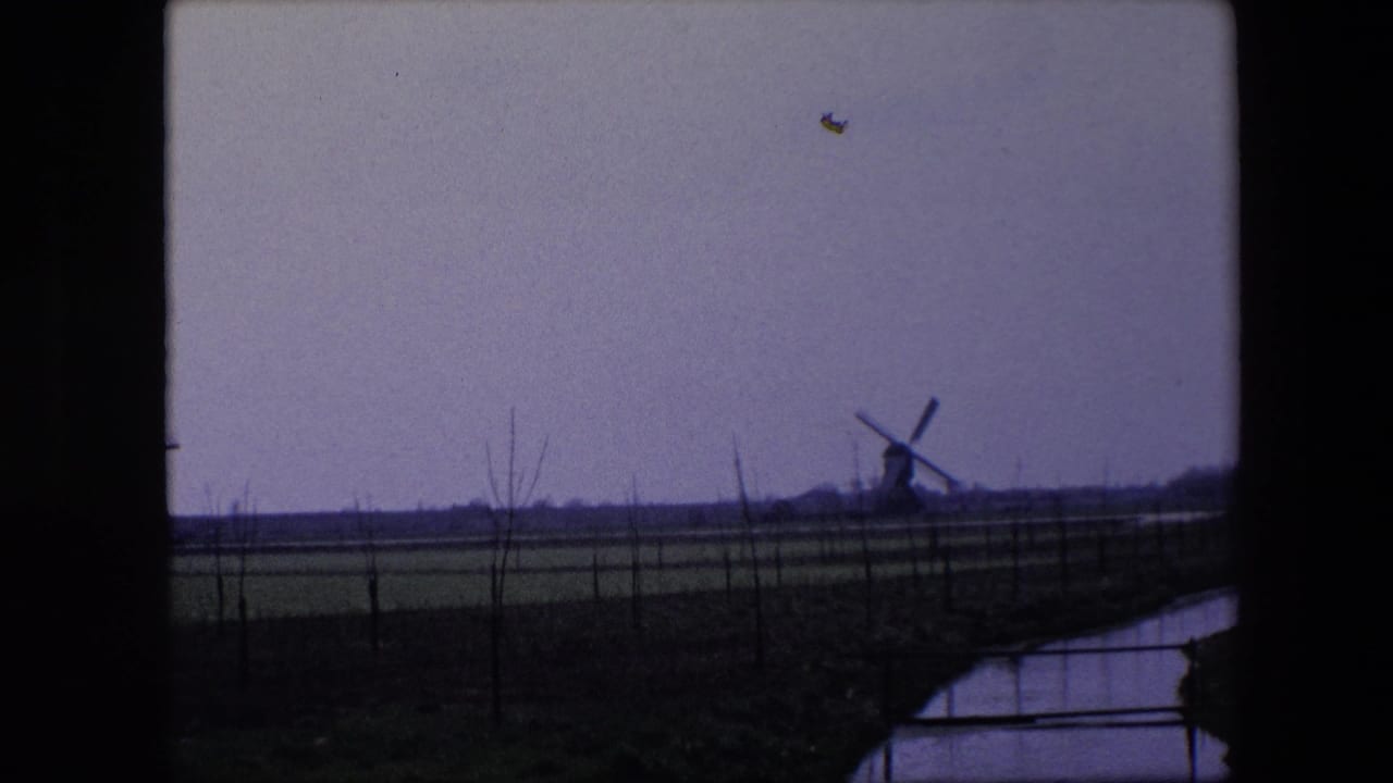 Final 1967 Belgium home-movie frame showing a windmill on the horizon across flat farmland, with fence posts, bare vegetation, and a narrow canal in the foreground.