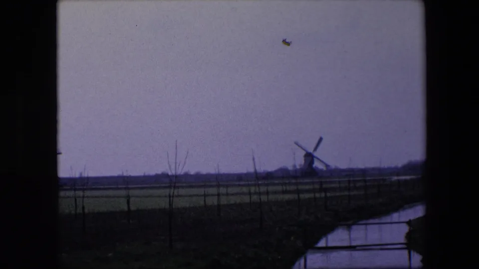 Final 1967 Belgium home-movie frame showing a windmill on the horizon across flat farmland, with fence posts, bare vegetation, and a narrow canal in the foreground.