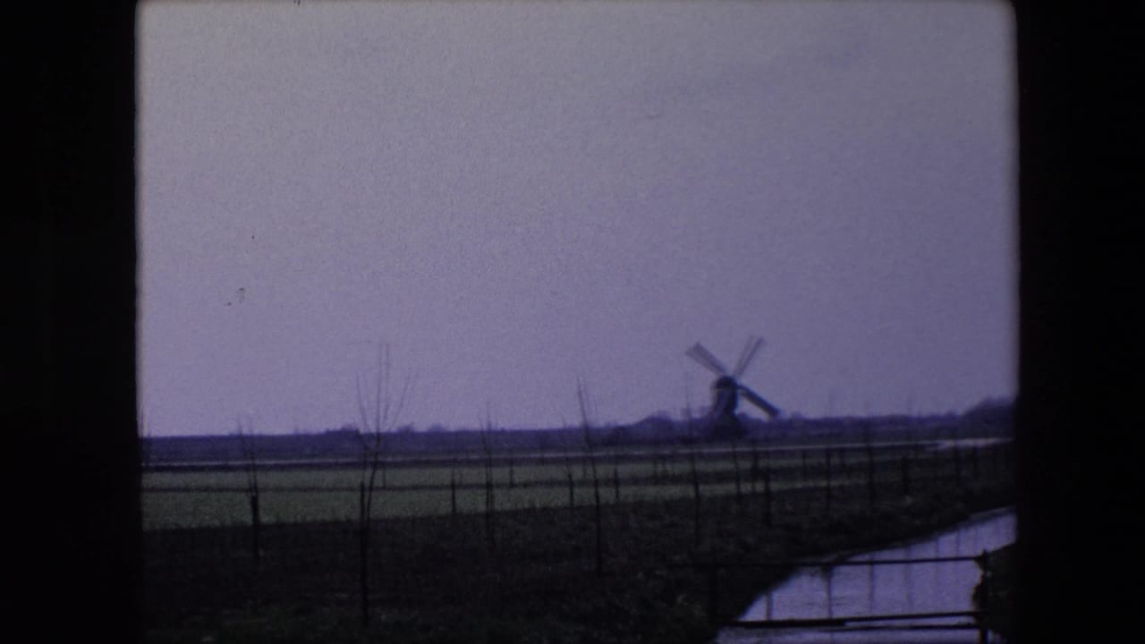 1967 8mm home-movie frame near Brussels showing a distant windmill across flat fields, with a narrow canal and dark window framing in the foreground.
