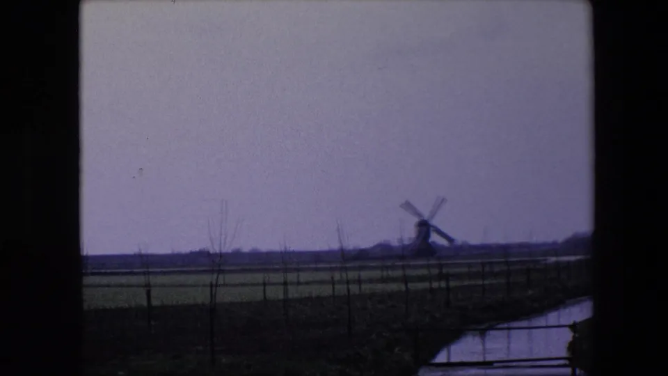 1967 8mm home-movie frame near Brussels showing a distant windmill across flat fields, with a narrow canal and dark window framing in the foreground.