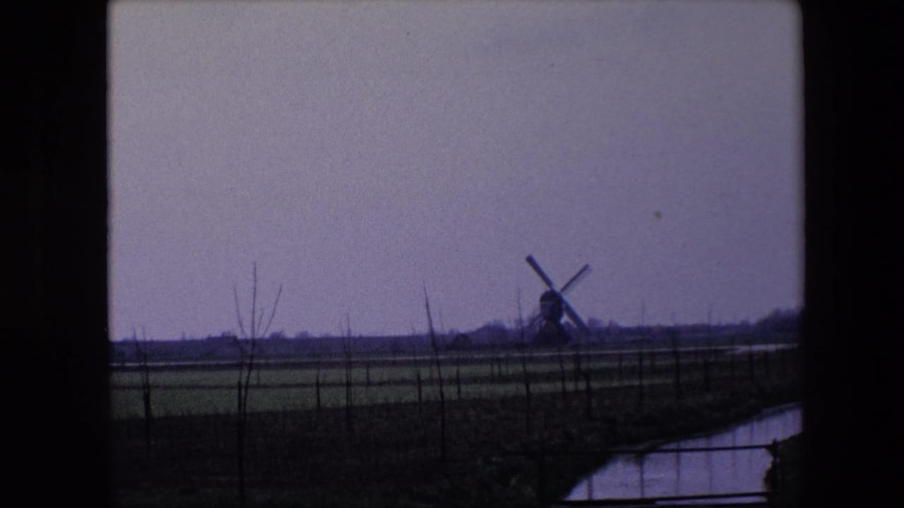 1967 Brussels-area frame with a far windmill standing beyond open fields, bare tree trunks, and a canal under a pale overcast sky.