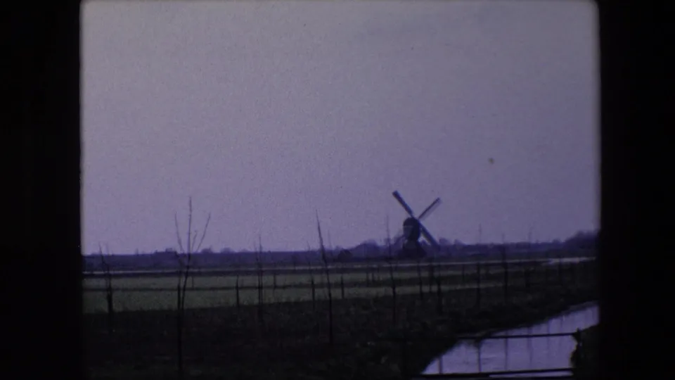 1967 Brussels-area frame with a far windmill standing beyond open fields, bare tree trunks, and a canal under a pale overcast sky.