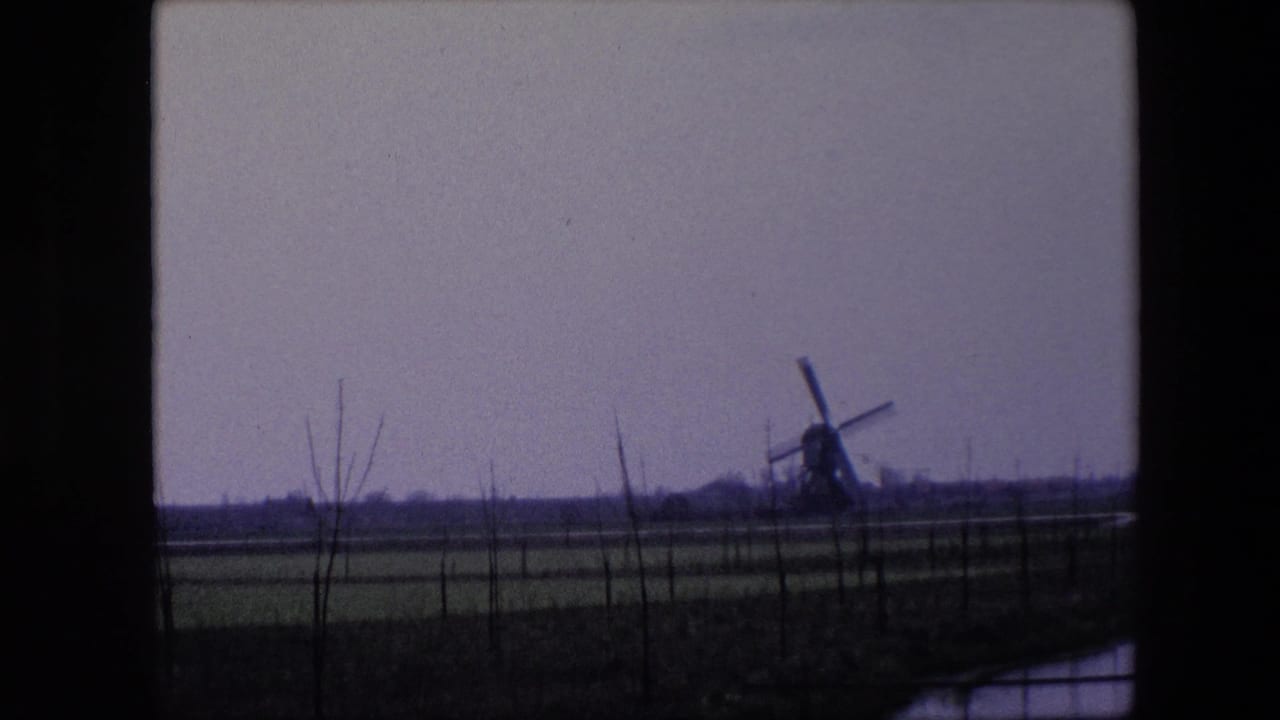 8mm frame from 1967 near Brussels showing a distant windmill, low agricultural fields, sparse trees, and dark window or vehicle framing.
