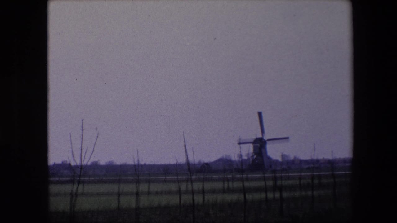 1967 Brussels-area home-movie frame with the windmill more centered in the distance, surrounded by flat fields, sparse trees, and a muted overcast sky.