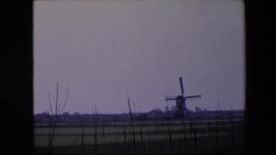 1967 Brussels-area home-movie frame with the windmill more centered in the distance, surrounded by flat fields, sparse trees, and a muted overcast sky.
