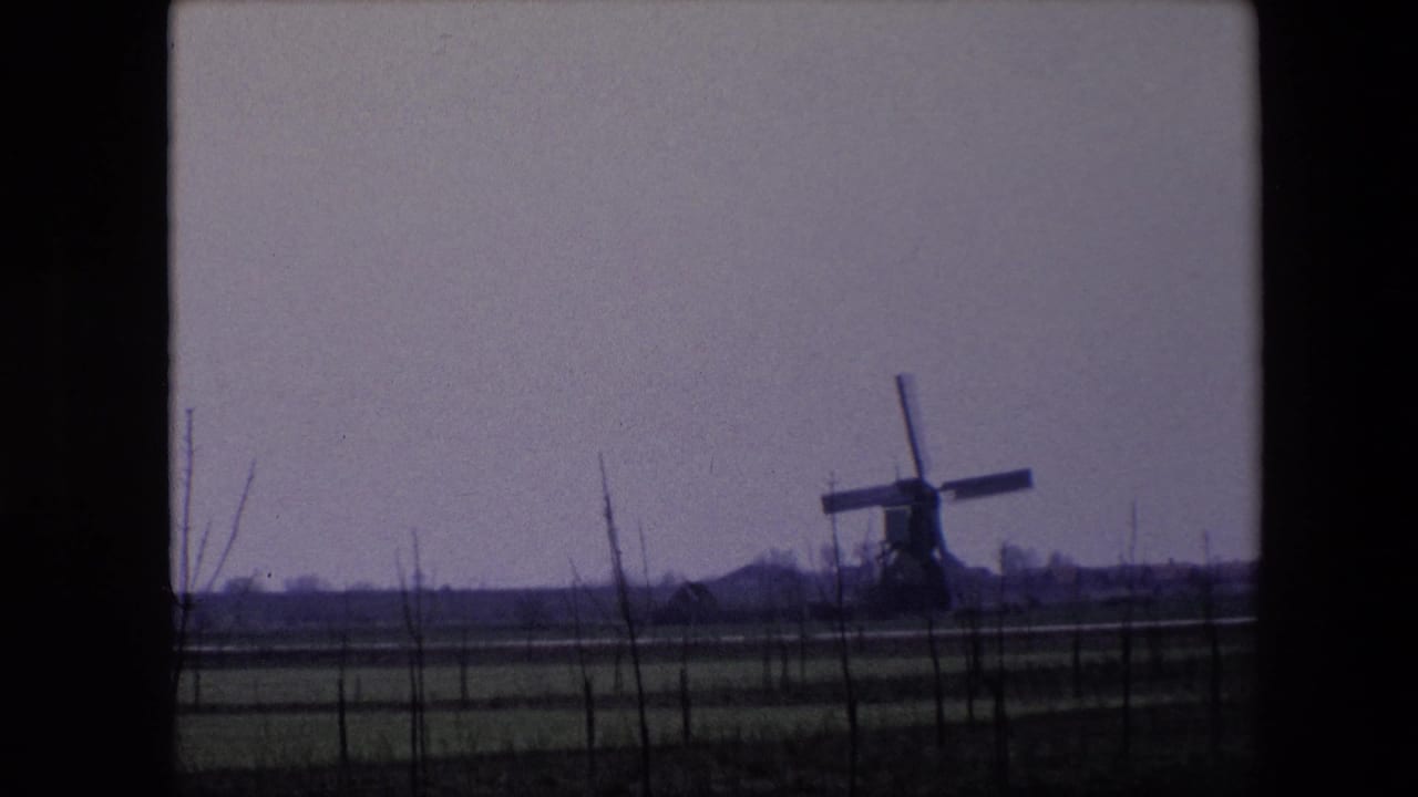 1967 Brussels-area 8mm frame showing a traditional windmill across flat fenced farmland, with bare branches in the foreground and a muted sky.