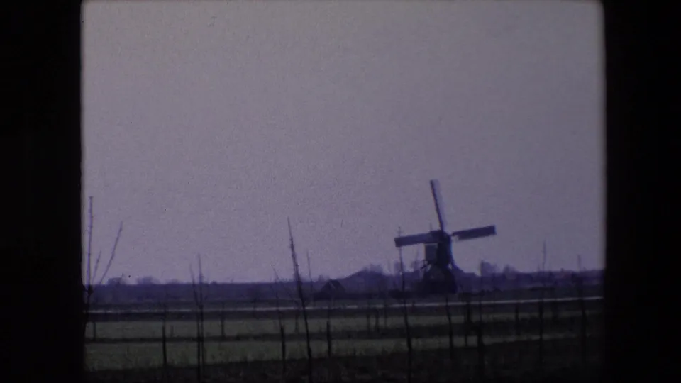 1967 Brussels-area 8mm frame showing a traditional windmill across flat fenced farmland, with bare branches in the foreground and a muted sky.