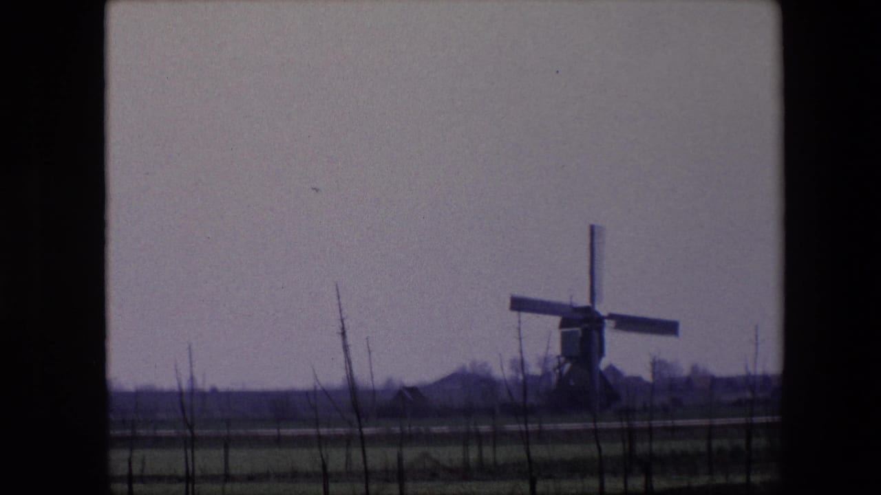 1967 Brussels-area 8mm frame with the windmill centered beyond open fields, thin foreground branches, and distant low structures on the horizon.
