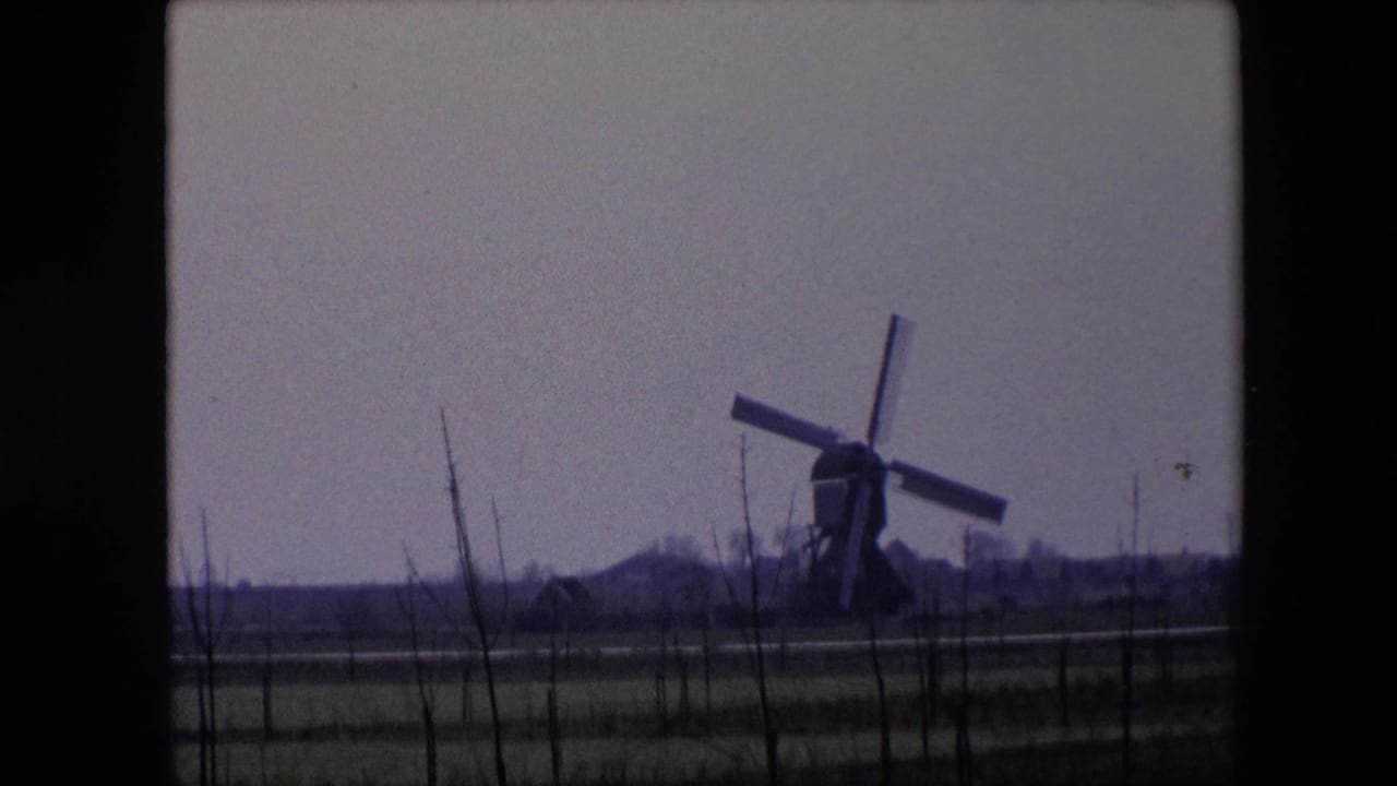 1967 Brussels-area 8mm frame showing the windmill sails at a new angle, with fenced fields, bare saplings, and distant rural buildings visible.