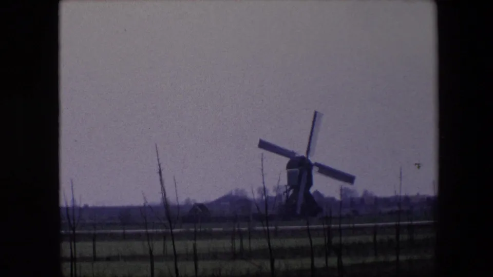 1967 Brussels-area 8mm frame showing the windmill sails at a new angle, with fenced fields, bare saplings, and distant rural buildings visible.